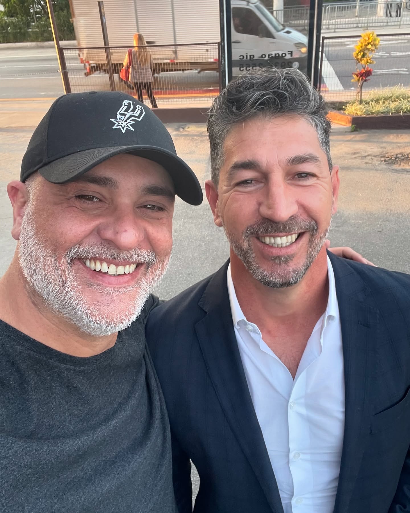 Selfie recente de Marcos, de boné preto, e Sérgio, de blazer e camisa branca, sorrindo abraçados na rua